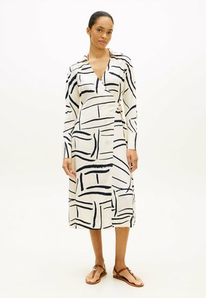 Mujer de pie vistiendo un vestido midi cruzado blanco con líneas abstractas negras y sandalias planas marrones sobre un fondo liso y claro.