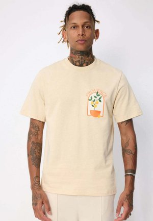 Homme avec des tatouages portant un t-shirt beige avec un graphique d’un citronnier en pot et le texte « PROVENCE » sur la partie supérieure gauche de la poitrine.