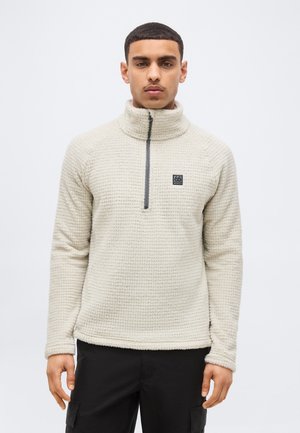 Jeune homme portant un pull en polaire texturé beige avec une fermeture éclair partielle et un pantalon noir, debout devant un fond clair uni.