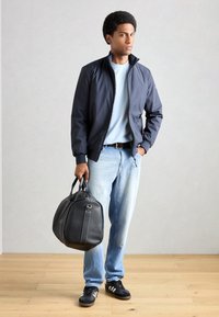 Marine bomberjakke, lyseblå skjorte, lyseblå jeans, sort bælte, holder en sort lædertaske med hvid syning, sorte sneakers.