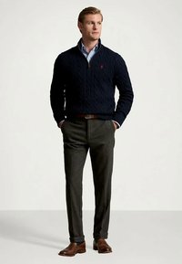 Mann som står med hendene i lommene, iført marineblå kabelstrikket genser, lyseblå skjorte, mørke bukser og brune lær sko.