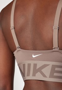 Nike sportski grudnjak u taupe nijansi s podesivim trakama, s širokim trakama s logotipom i malim bijelim swooshom na stražnjoj strani. Glatki, rastezljivi materijal.