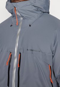 Šedá nepromokavá bunda s nastavitelnou kapucí, stříbrným zipem a kontrastními oranžovými šňůrkami. Má dvě přední kapsy na zip.