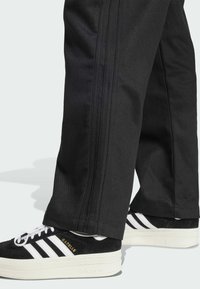 Pantalones negros con tres rayas verticales en los costados, combinados con zapatillas Adidas negras y blancas que presentan una parte superior de ante texturizado y suela de plataforma.
