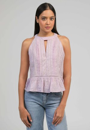 Femme portant un haut sans manches lavande avec motif ajouré, ourlet à basque et col montant, associée à un jean bleu clair, debout devant un fond gris.