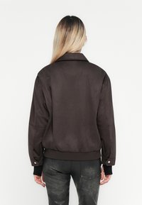 Bruine bomberjack met een gladde textuur, oversized pasvorm en een kraag. Voorzien van ribgebreide manchetten en een tailleband. Draagt met donkere metallic jeans.