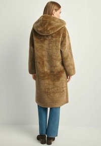 Brązowy płaszcz z futra ekologicznego z kapturem, o luźnym kroju i długich rękawach, zestawiony z jasnoniebieskimi dżinsami i ciemnobrązowymi botkami za kostkę.