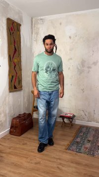 Άντρας με dreadlocks στέκεται σε ένα δωμάτιο με φθαρμένους μπεζ τοίχους, φορώντας ένα πράσινο γραφικό T-shirt, μπλε τζιν και μαύρα παπούτσια, κοντά σε ένα δερμάτινο σακίδιο και ένα μοτίβωτο χαλί.