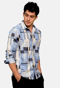 Camicia con bottoni caratterizzata da un motivo a scacchi astratto in blu chiaro, bianco e nero, maniche lunghe arrotolate fino a metà avambraccio e colletto classico.