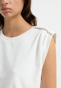 Witte top met korte mouwen, gemaakt van een zachte stof, met een ronde halslijn en parelversieringen op de schouder voor extra detail.