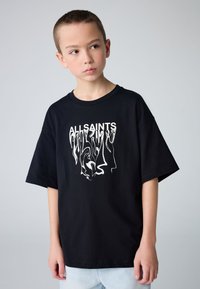 Poiss, kellel on lühikesed juuksed, kannab musta, üle mõõdu suure suurusega T-särki, millel on valge moonutatud "ALLSAINTS" kiri, ja helesiniseid teksaseid, halli tausta ees.