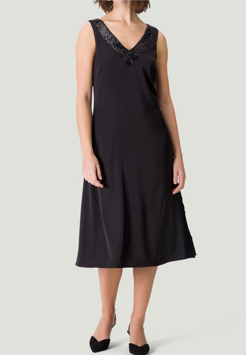 Femme portant une robe midi noire sans manches avec un décolleté en V pailleté et des escarpins noirs à bouts pointus, debout devant un fond uni.