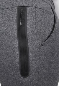 Šedé látkové kalhoty s hladkou texturou, vybavené zipovou kapsou po straně s černými akcenty a kontrastním černým lemováním zipu.