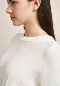 Weißer Strickpullover mit geripptem Struktur und rundem Ausschnitt, auf dem vertikale Streifenmuster im Stoff sichtbar sind.