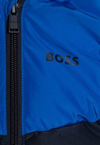 Doudoune bleue avec une fermeture éclair noire, un logo prononcé et une texture lisse et brillante. L'ourlet inférieur est noir.