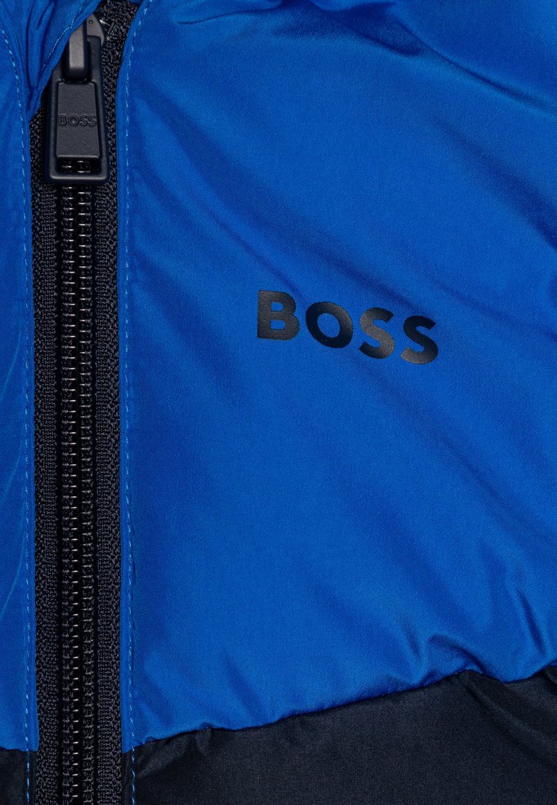 Doudoune bleue avec une fermeture éclair noire, un logo prononcé et une texture lisse et brillante. L'ourlet inférieur est noir.