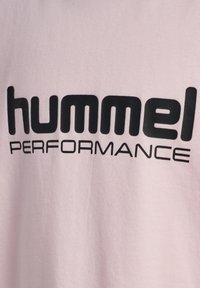 Jasnoróżowy t-shirt z wyraźnym czarnym nadrukiem, który głosi "hummel PERFORMANCE" na klatce piersiowej. Miękka tkanina o gładkiej fakturze.
