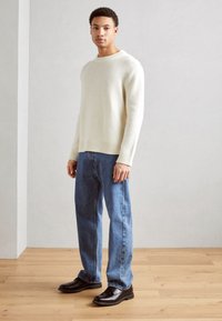 Pulover tricotat de culoare crem, cu manșete și tiv striate, combinat cu blugi drepti de culoare albastră și pantofi negri slip-on. Fundal din lemn.