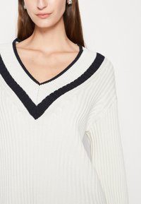 Kvinna som bär en vit ribbad tröja med en svart dubbelfärgad V-ringning och silverörhängen, sedd från bröstet och upp.