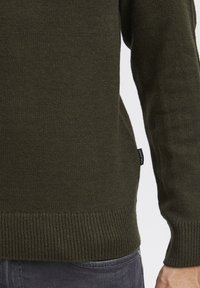 Pulover verde khaki din țesătură tricotată texturată. Are manșete și tiv cu nervuri, plus un mic etichetă a mărcii pe lateral.