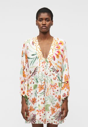 Nő, aki hosszú ujjú, V-nyakú ruhát visel, amely színes, narancssárga, zöld és rózsaszín virágmintával van díszítve fehér alapon, sima háttér előtt áll.