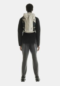 Sac à dos avec un corps en tissu beige, des panneaux latéraux noirs et des accessoires en argent. Il dispose de bretelles réglables et d'une fermeture éclair sur le dessus.