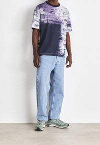 Krátký rukáv tričko s abstraktním vzorem v purpurové a námořnické modré barvě, spojené se světle modrými džínami a zelenými sportovními teniskami.