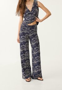 Vrouw in een marineblauwe mouwloze top met bloemenprint en bijpassende wijde broek, staande met één hand op haar heup en de andere hand omhoog om de top op te tillen.