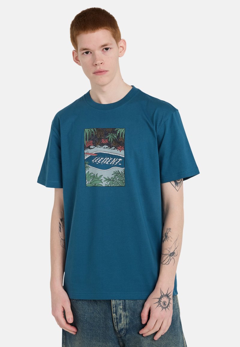 Giovane uomo con capelli rossi indossa una maglietta blu con una grafica colorata di una piscina tropicale, abbinata a jeans in denim scolorito.