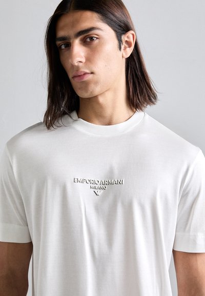 Young man with shoulder-length dark hair wears a plain white t-shirt with "EMPORIO ARMANI MILANO" logo on the chest against a gray background.