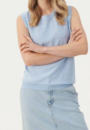 Femme aux cheveux blonds ondulés porte un haut bleu clair sans manches et une jupe en jean claire, les bras croisés, face à l'avant sur fond uni.