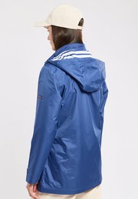 Giacca impermeabile blu con cappuccio, caratterizzata da strisce bianche sulla parte posteriore e maniche regolabili. Tessuto liscio e lucido.