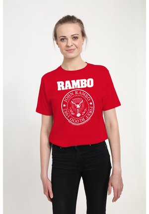 Ung kvinde med lyst hår bundet tilbage iført en rød t-shirt med hvid Rambo-tekst og emblem, kombineret med sorte bukser, stående på hvid baggrund.