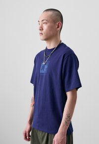T-shirt en coton bleu marine à manches courtes avec un motif graphique d'ours. Le modèle porte des colliers en chaîne argentée superposés.