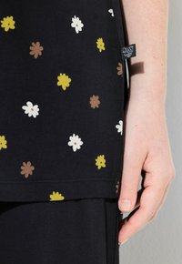Túnica negra de tela con un patrón de pequeñas flores multicolores en amarillo, blanco, marrón y verde. Etiqueta de tela visible en el lateral.