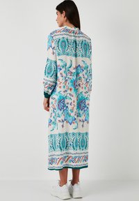 Long, floral-print dress in white with blue and pink patterns. Features long sleeves and a straight silhouette, paired with white sneakers.