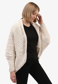 Vrouw met kort blond haar, gekleed in een crèmekleurige gebreide cardigan met textuur over een zwart shirt en zwarte broek, die naar beneden kijkt met een hand dichtbij haar hoofd.