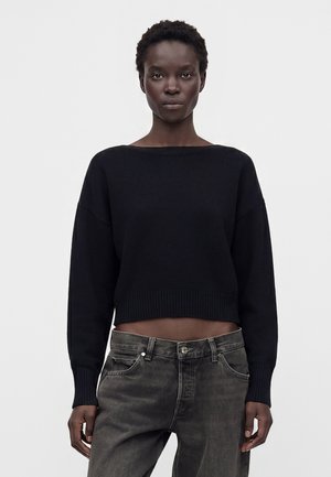 Sort person iført en sort langærmet cropped sweater og falmede sorte jeans, stående mod en ensfarvet hvid baggrund.