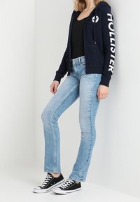 Felpa zip-up blu navy con la scritta "Hollister" stampata in bianco, abbinata a jeans dritti azzurri e sneakers in tela nere.