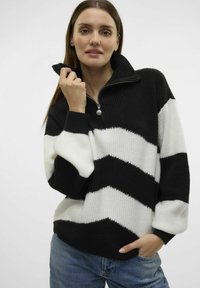 Sweter w czarno-białą zygzakowatą kratę z wysokim kołnierzem, prążkowaną strukturą i zamkiem błyskawicznym, zestawiony z niebieskimi dżinsami.