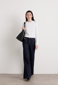 Una donna con lunghi capelli scuri cammina con sicurezza, indossando una maglietta bianca aderente e jeans scuri a gamba larga, mentre porta una borsa nera.