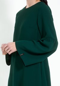 Zelena haljina dugih rukava s okruglim izrezom i suptilnim nabranjima na manžetama. Izrađena od glatkog materijala s detaljem bočne džepne.