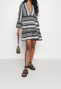 Svart och vit mönstrad klänning med långa ärmar, V-hals, samlad midja, kombinerad med svarta sandaler och en olivgrön handväska.