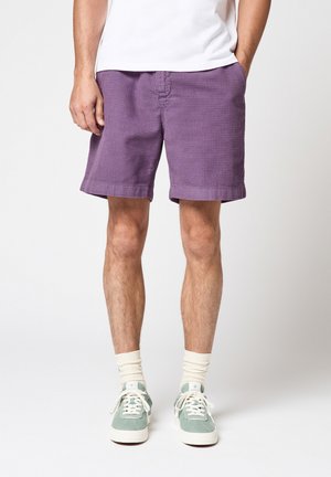 Homme portant un short violet texturé, des chaussettes blanches, des baskets vert pastel et une chemise blanche, debout devant un fond uni.