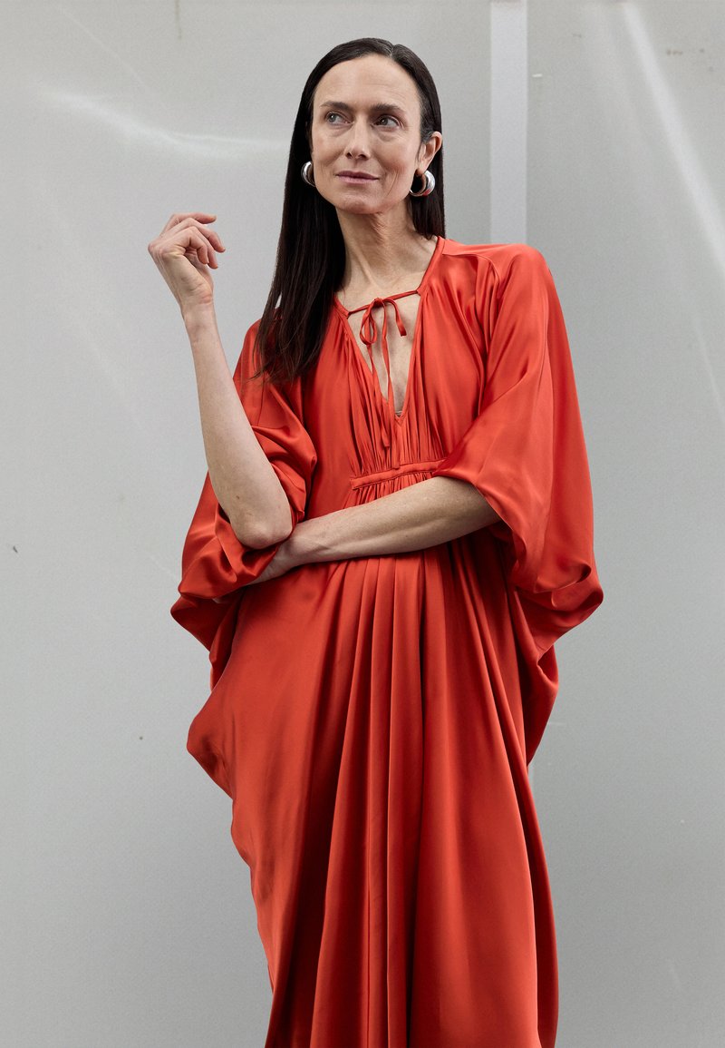 Vestido de satén rojo con mangas anchas y drapeadas, escote profundo con cordones para atar y pliegues verticales desde la cintura hacia abajo.
