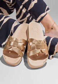 Gold slide sandals with a woven design, featuring a smooth leather texture and a light tan insole. Accented with a flower logo imprint.