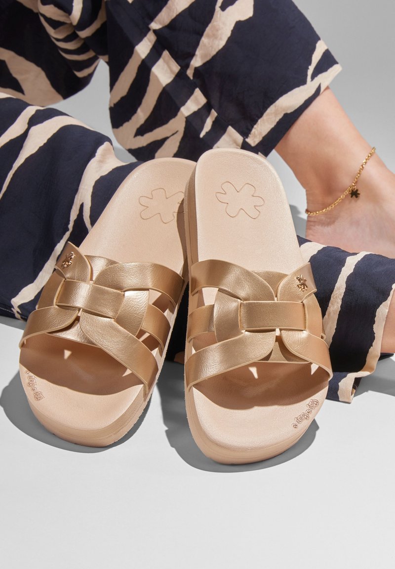 Gold slide sandals with a woven design, featuring a smooth leather texture and a light tan insole. Accented with a flower logo imprint.