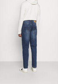 Dark blue denim jeans with a relaxed fit, five pockets, and a light brown leather patch at the back. Worn with a white hoodie and sneakers.