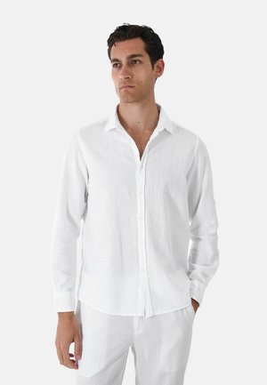 Uomo con capelli scuri che indossa una camicia bianca a maniche lunghe con bottoni e pantaloni bianchi, in piedi con una mano in tasca davanti a uno sfondo semplice.
