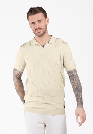 Hombre con tatuajes que lleva una camisa de polo beige de manga corta texturada y pantalones de color claro, de pie contra un fondo blanco.